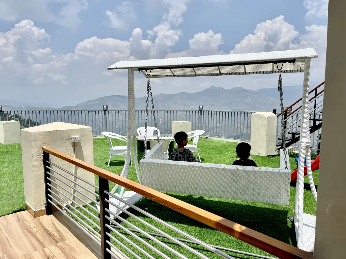 The rooftop lawn: a white swing with children on it, white chairs and a table, and the Himalaya beyond the railing.